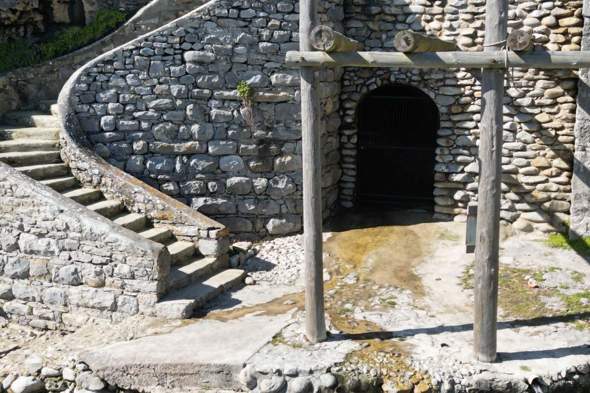 A rustic stone-built structure featuring a curved staircase and a gated entrance, set within a natural landscape with integrated stone masonry.