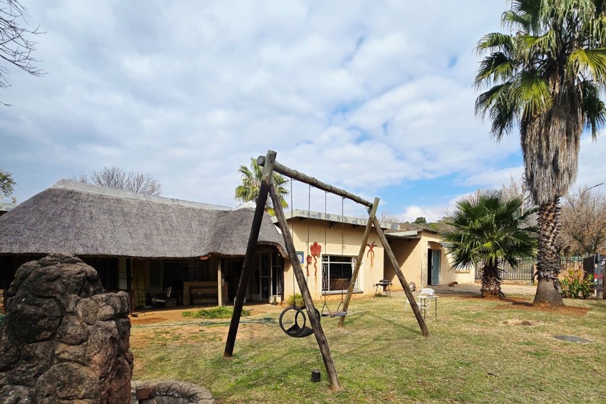 A spacious rear garden featuring a thatch-roofed dwelling, a stone water feature, and established palm trees with a large lawn area.