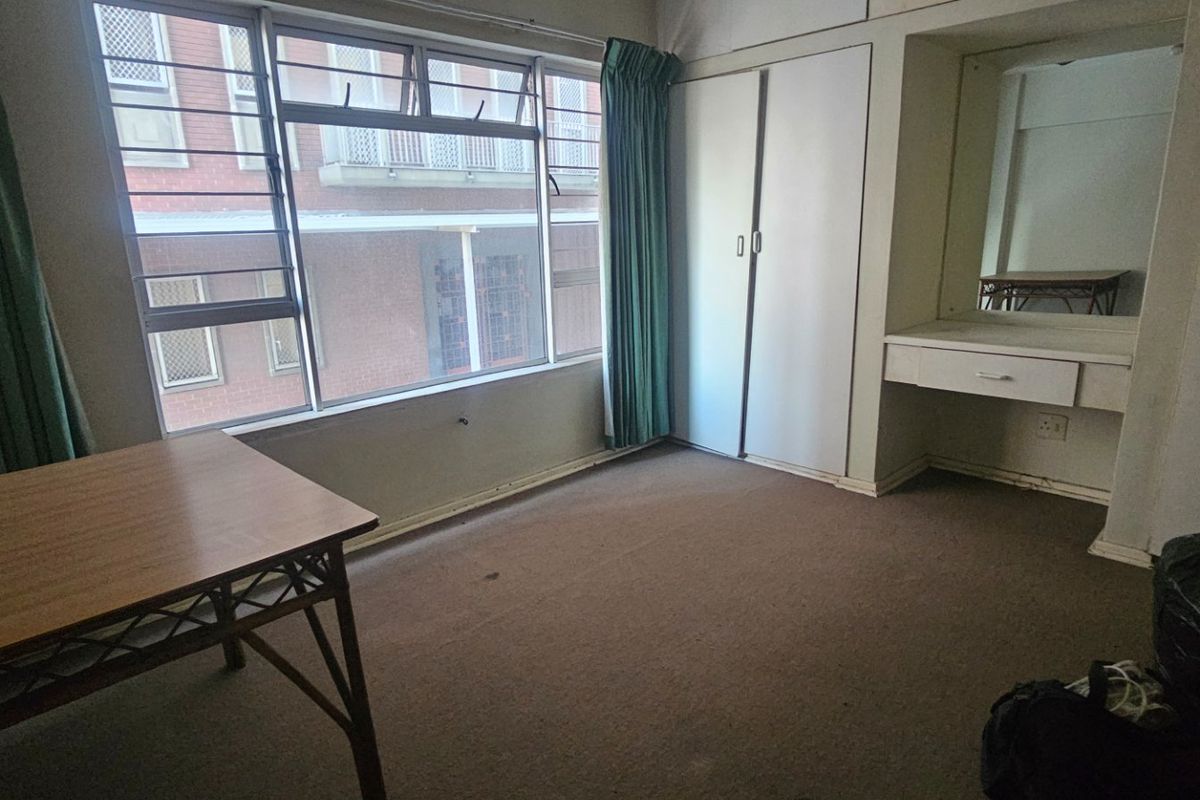 A bedroom featuring a large window, built-in wardrobe, and a built-in vanity unit, offering a functional space ready for renovation.