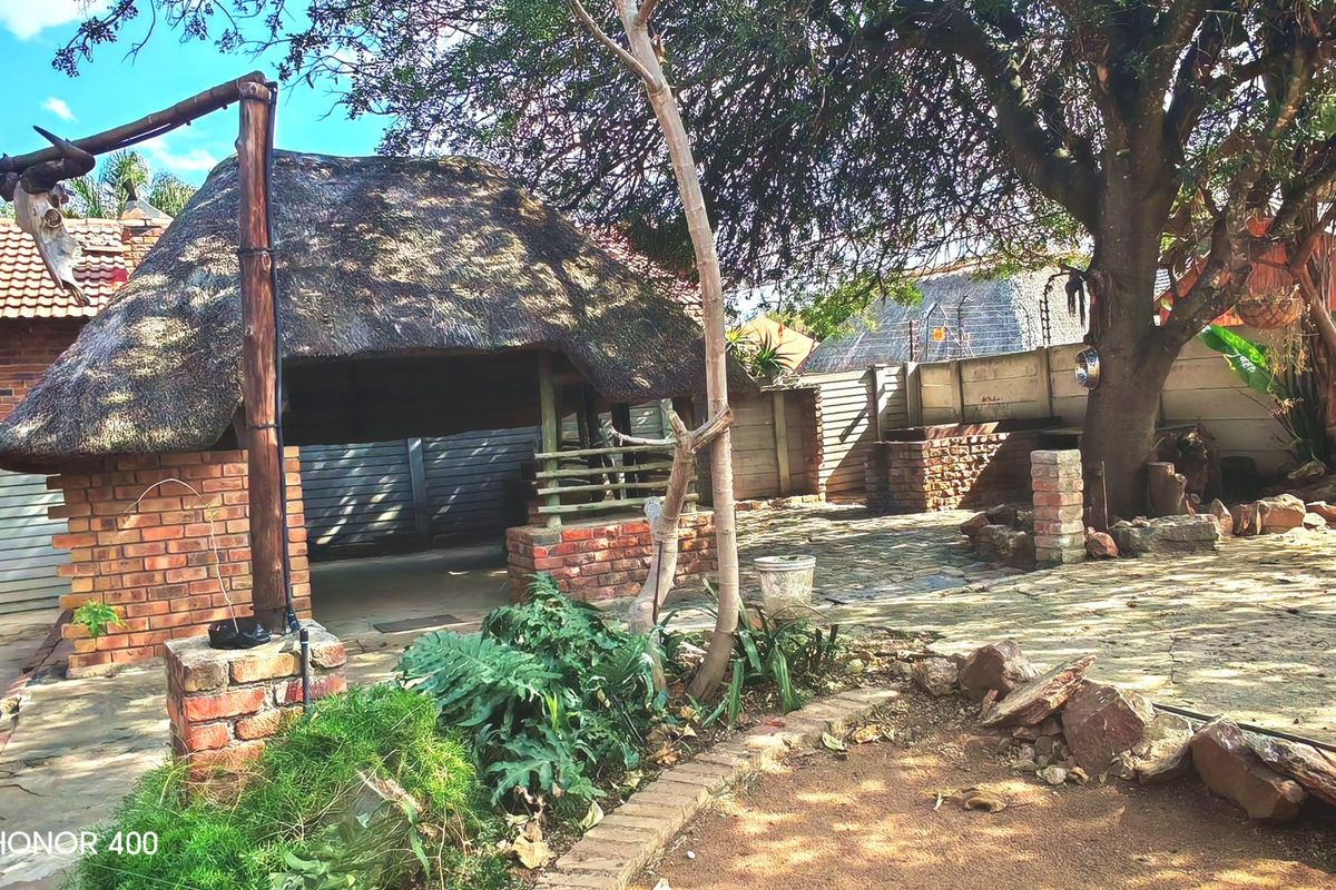 Rear garden area featuring a traditional thatch lapa, built-in brick braai, and paved patio space enclosed by a boundary wall with electric fencing.