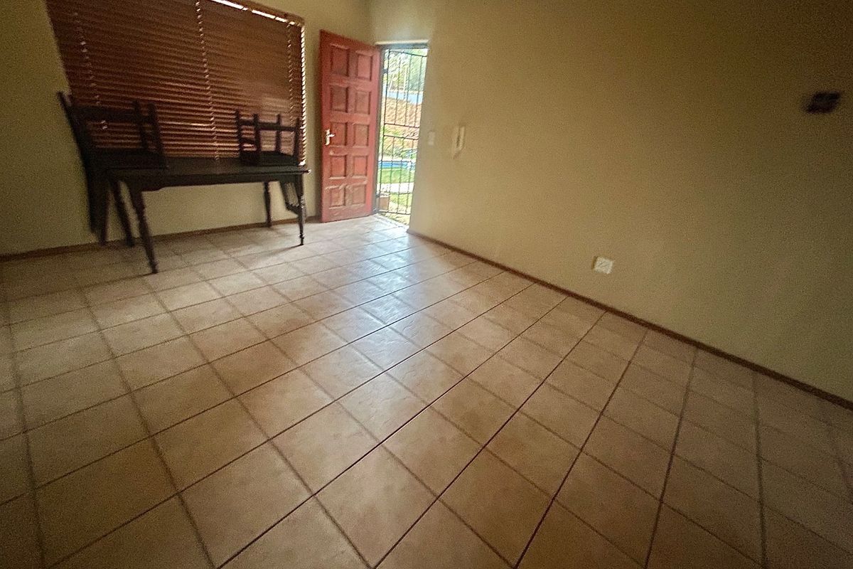 A spacious, empty living area featuring neutral tiled flooring, wooden blinds, and direct access to the exterior through a timber door with a security gate.