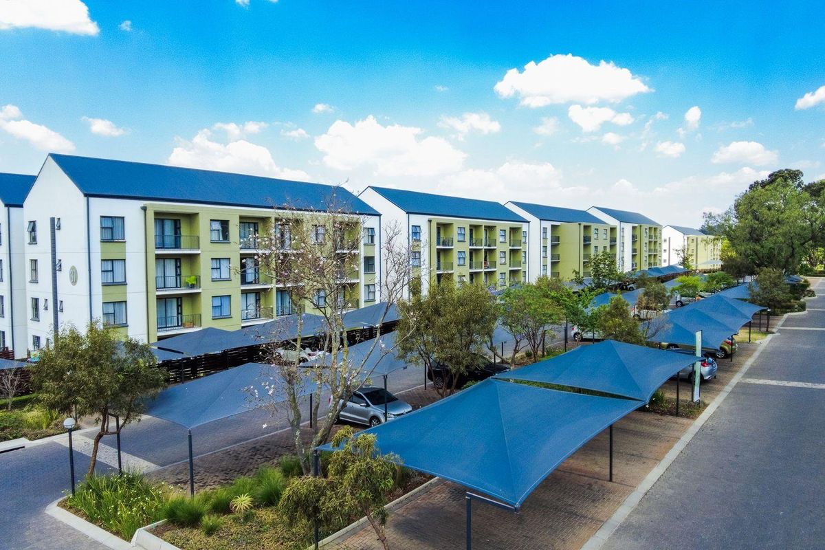 Aerial view of a modern apartment complex featuring multi-story residential buildings, paved parking areas with shade cloth carports, and landscaped grounds.