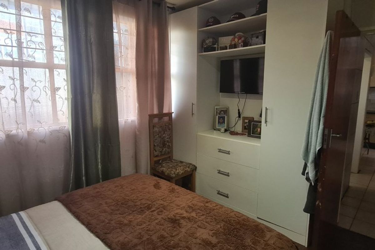 A bedroom featuring a built-in wardrobe with integrated shelving and security bars on the window, with a view into the adjacent kitchen area.