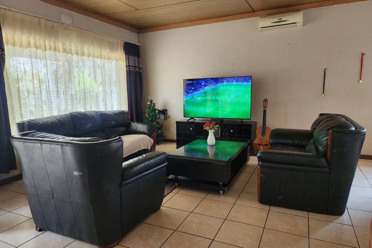 A spacious living room featuring tiled flooring, a timber-paneled ceiling, and a wall-mounted air conditioning unit, with large windows providing natural light.