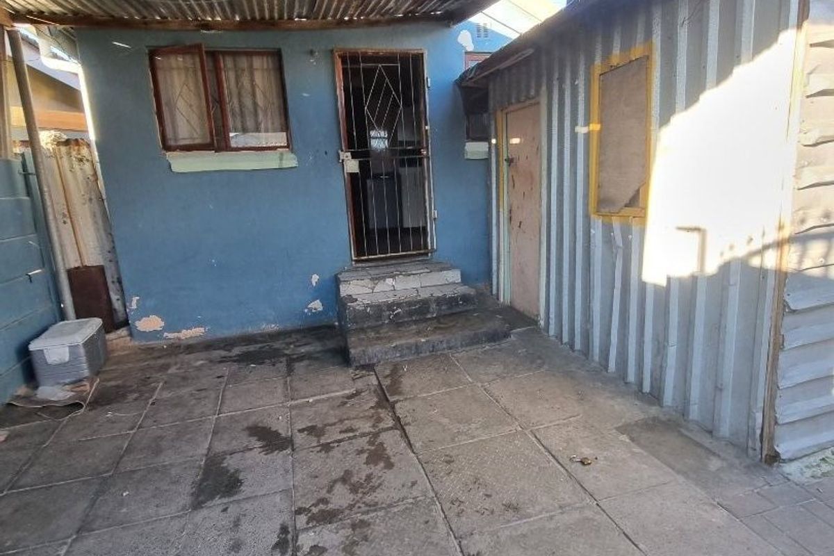 Rear exterior area featuring a paved patio, corrugated iron roofing, and metal wall cladding, showing significant signs of weathering and wear.