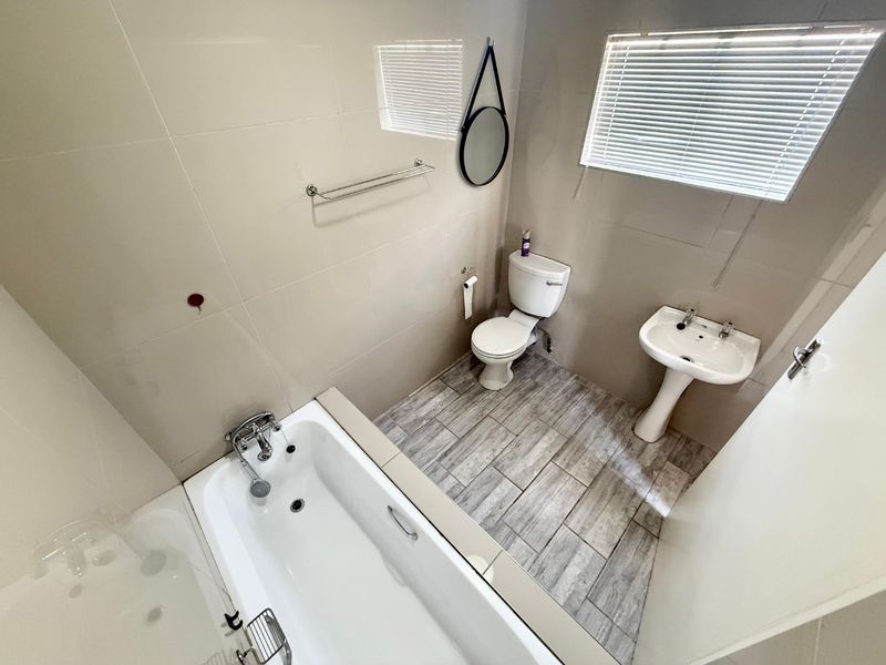 A clean, functional bathroom featuring a bathtub, pedestal basin, and toilet with neutral wall tiling and wood-look floor tiles.