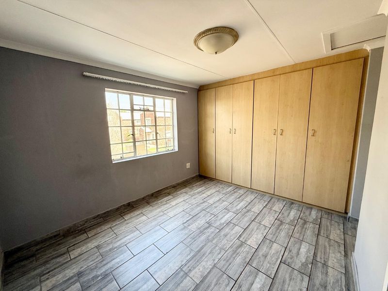 A bright, empty bedroom featuring modern wood-look tiled flooring, ample built-in cupboard storage, and a window with security bars.