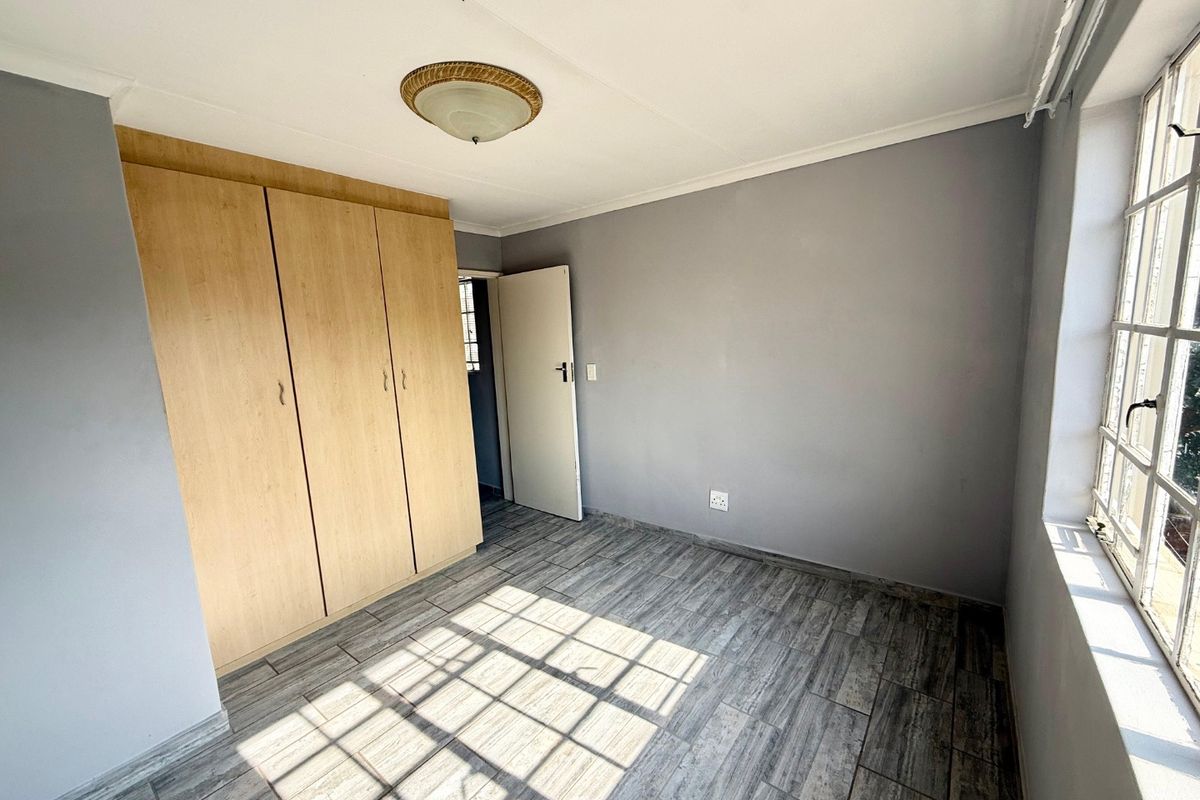 A bright, empty bedroom featuring modern wood-look tiled flooring, built-in cupboards, and a large steel-framed window allowing for ample natural light.