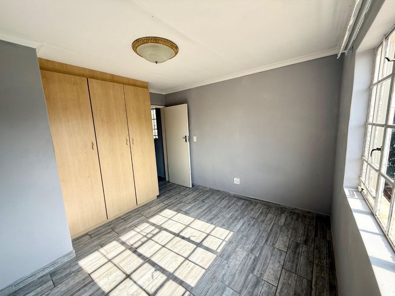 A bright, empty bedroom featuring modern wood-look tiled flooring, built-in cupboards, and a large steel-framed window allowing for ample natural light.