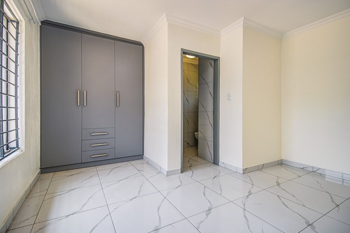 A bright, modern bedroom featuring sleek grey built-in cupboards, high-quality marble-look porcelain floor tiles, and an en-suite bathroom entrance.