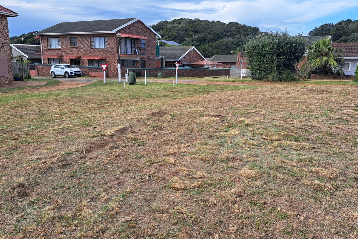 A view of a vacant residential plot featuring a grass lawn with surrounding residential properties in the background.