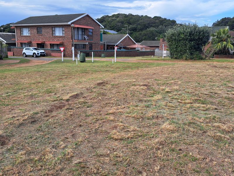 A view of a vacant residential plot featuring a grass lawn with surrounding residential properties in the background.
