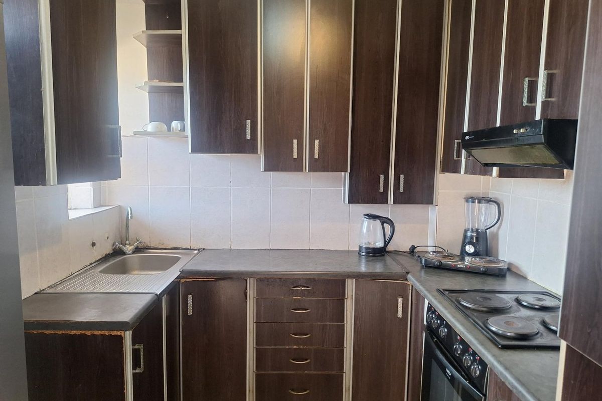 A compact kitchen featuring dark wood-finish cabinetry, laminate countertops, a built-in oven, electric hob, and a stainless steel sink with tiled backsplash.