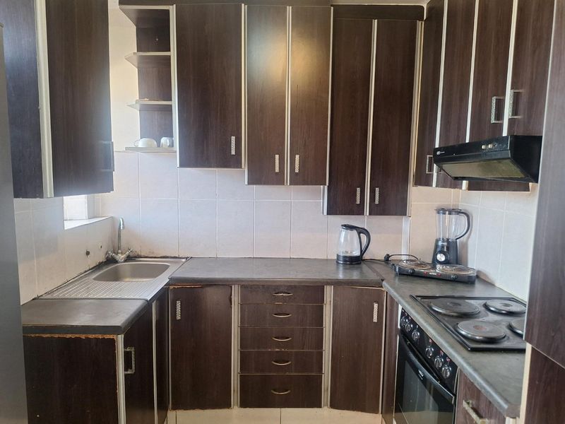 A compact kitchen featuring dark wood-finish cabinetry, laminate countertops, a built-in oven, electric hob, and a stainless steel sink with tiled backsplash.