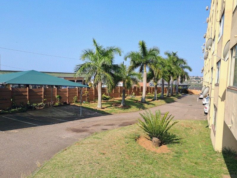 Exterior view of the apartment complex grounds featuring a paved driveway, covered parking bays, and established palm tree landscaping.