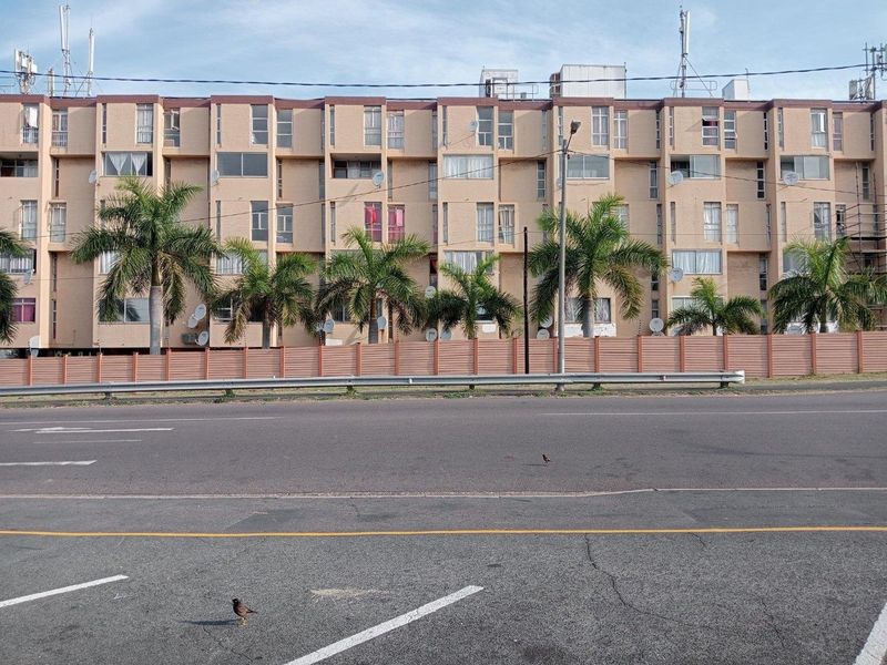Street-level view of a multi-story apartment complex featuring a brick facade, perimeter fencing, and mature palm trees along the front boundary.