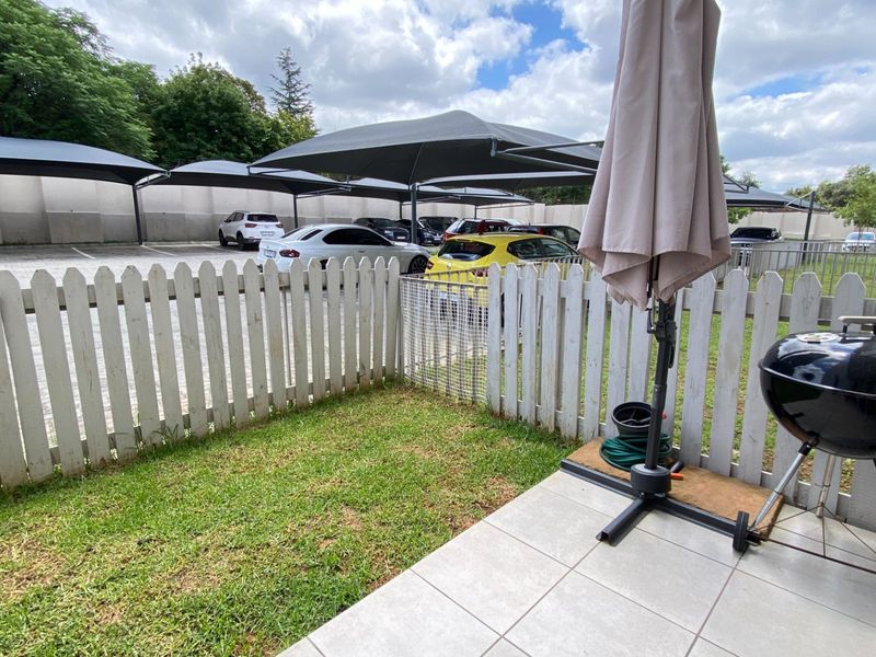 A private rear garden area featuring a paved patio, small lawn, and white picket fencing, overlooking a communal parking area with shade cloth carports.