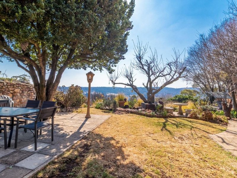A landscaped garden featuring a paved patio area, mature trees, and a stone retaining wall with scenic views.