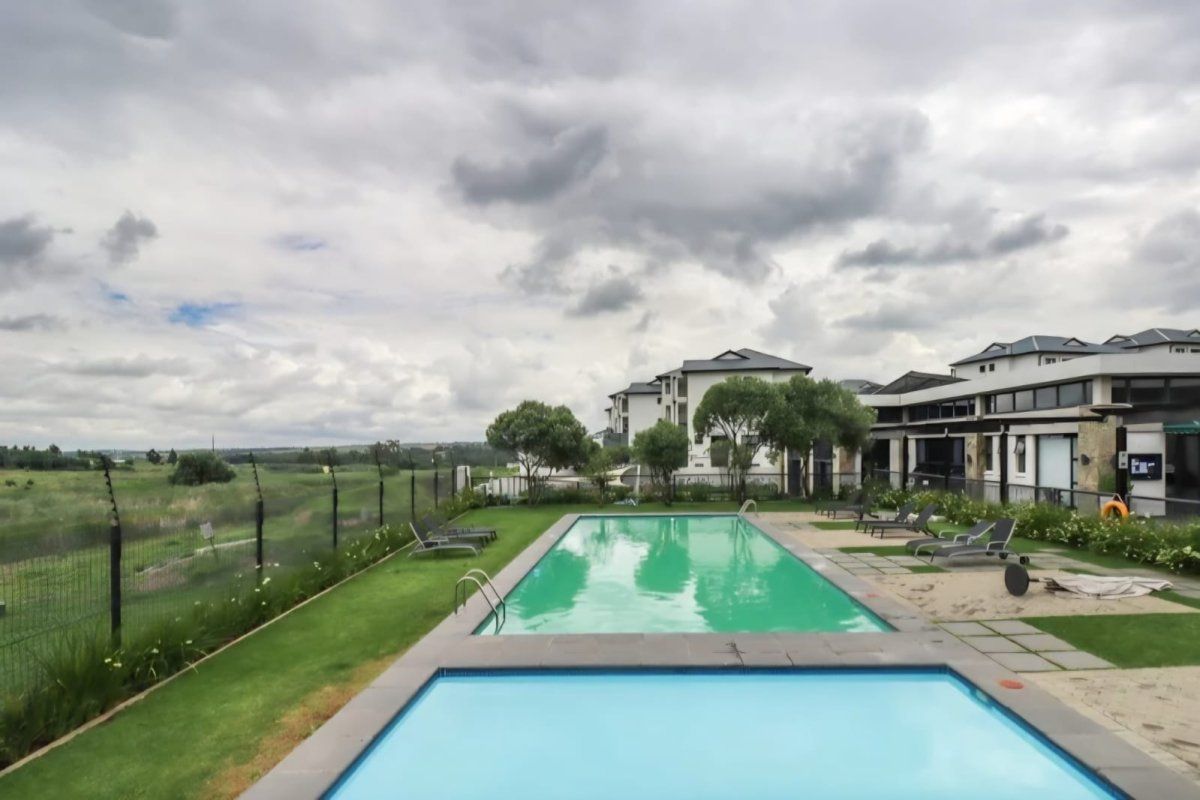 A well-maintained communal swimming pool area featuring paved surrounds, landscaped lawn, and perimeter fencing with views of the surrounding estate.