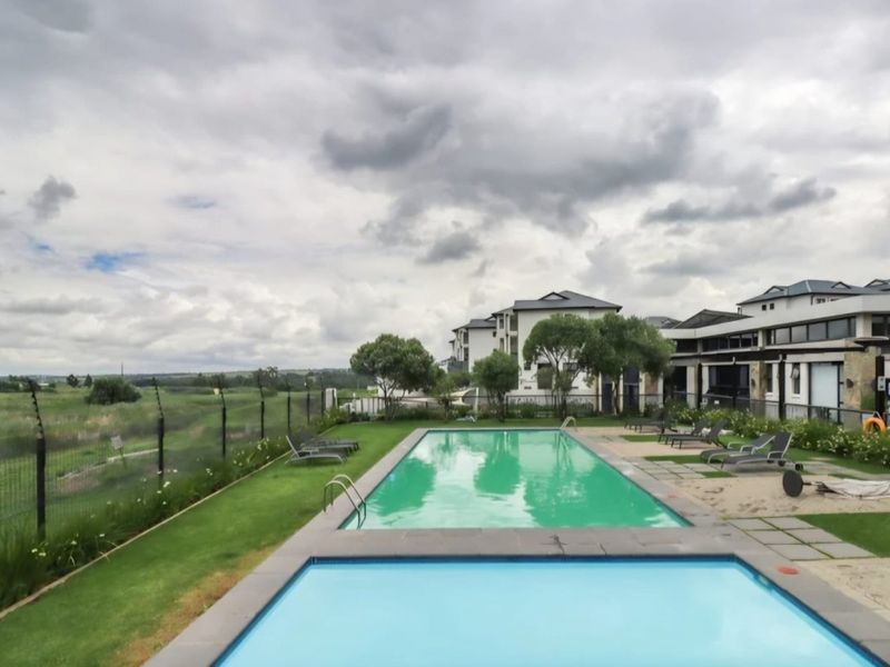 A well-maintained communal swimming pool area featuring paved surrounds, landscaped lawn, and perimeter fencing with views of the surrounding estate.