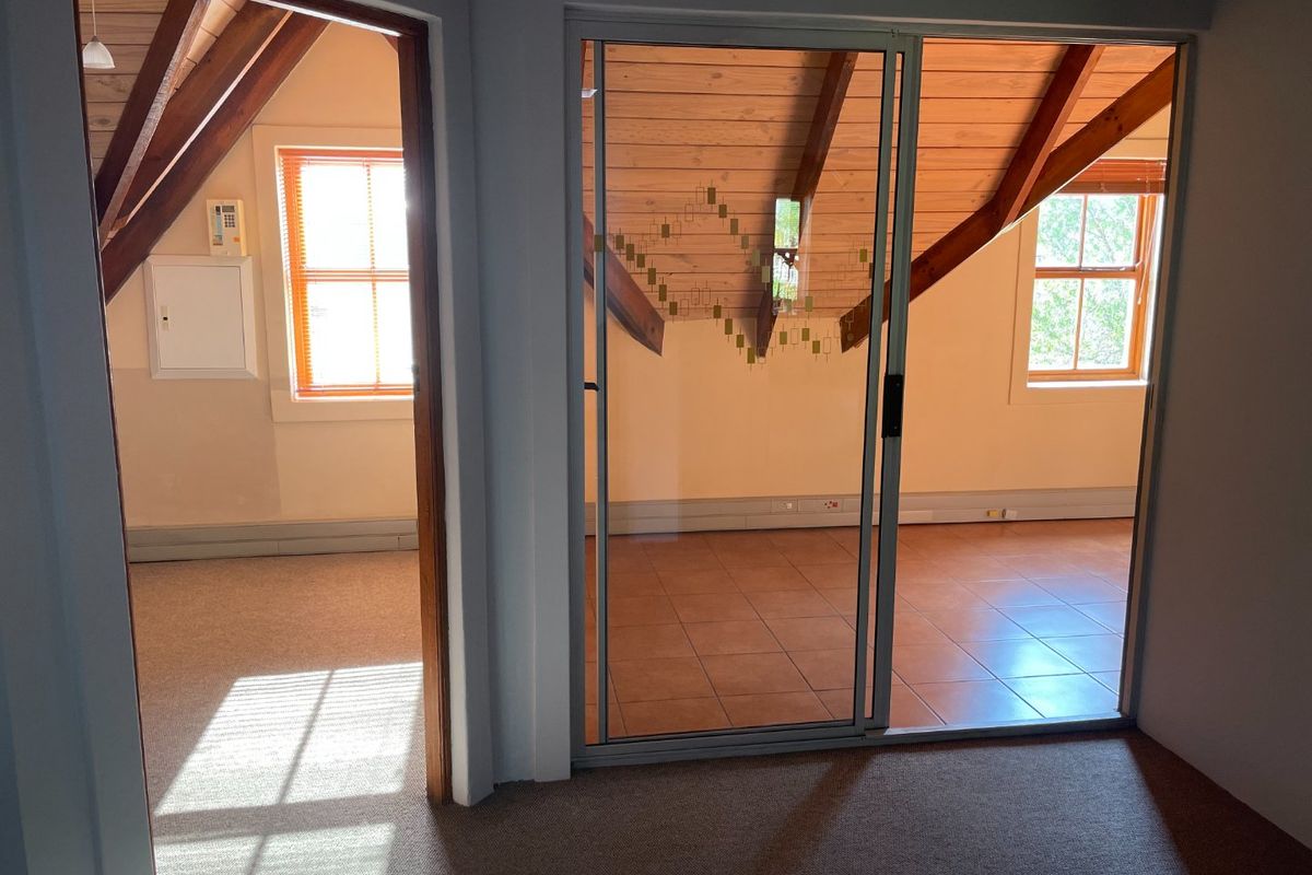An empty office space featuring exposed timber rafters, tiled and carpeted flooring, and a sliding glass door providing natural light.