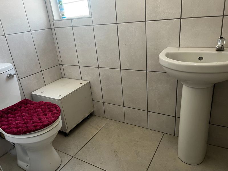 A clean, functional bathroom featuring a pedestal washbasin, standard toilet, and neutral ceramic wall and floor tiling.