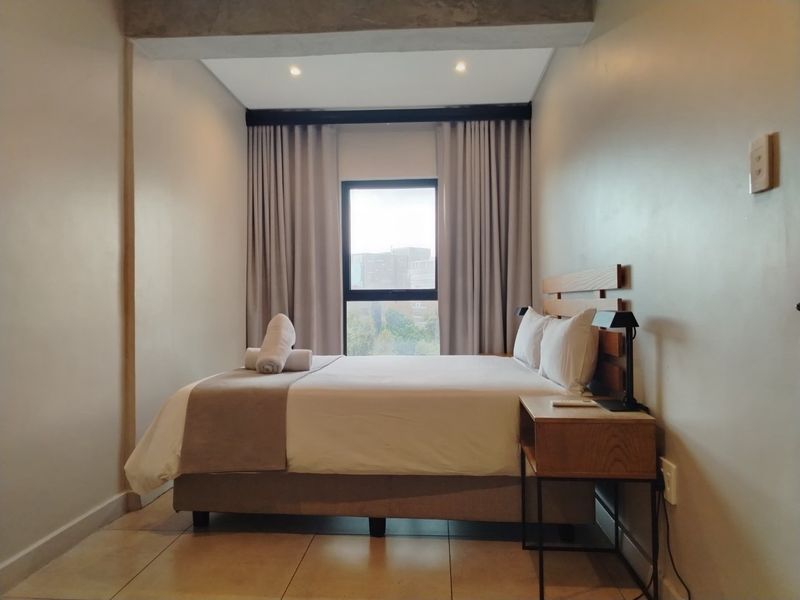 A well-maintained bedroom featuring neutral wall tones, tiled flooring, recessed ceiling lighting, and a window with integrated curtain tracks.