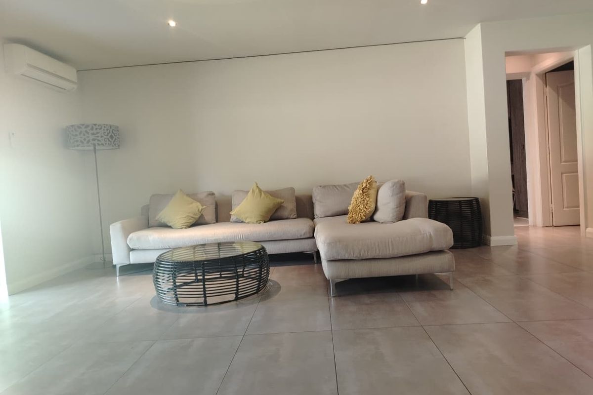 A bright and modern living room featuring large format floor tiles, recessed downlighting, and a wall-mounted split-system air conditioning unit.
