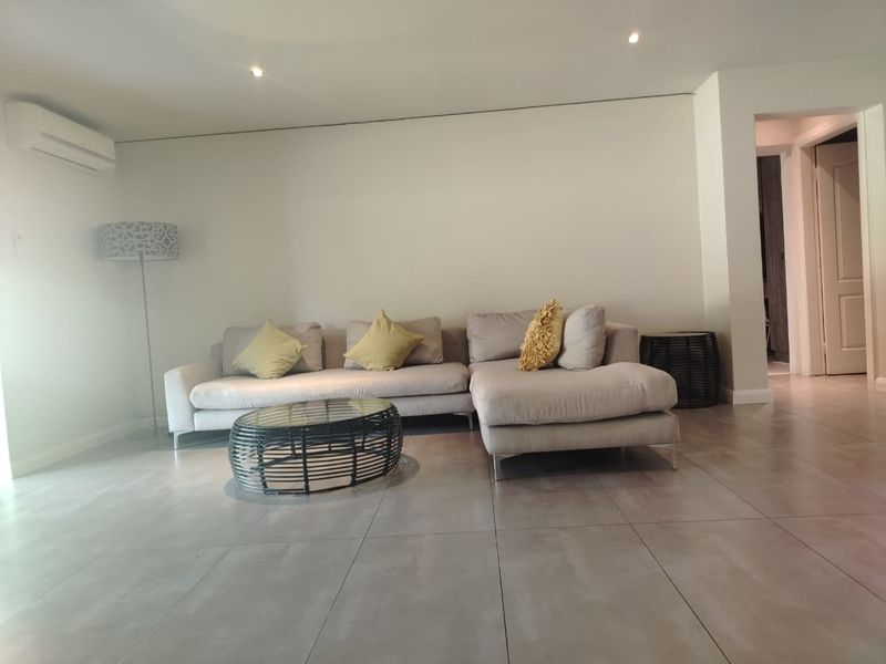 A bright and modern living room featuring large format floor tiles, recessed downlighting, and a wall-mounted split-system air conditioning unit.