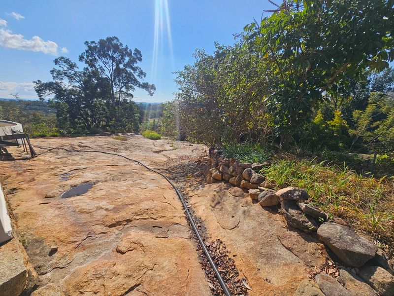 A natural outdoor garden area featuring a large rock outcrop, stone retaining wall, and established trees with scenic views.