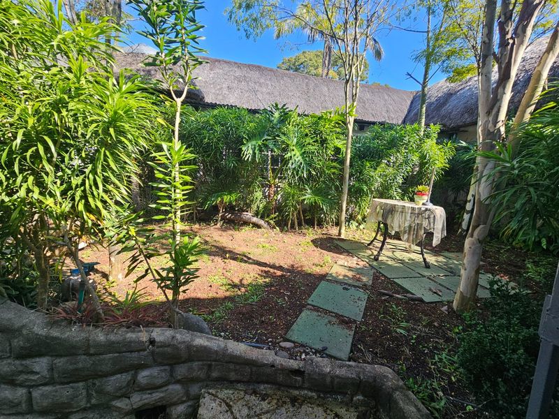 A private garden area featuring a stone retaining wall, paved walkway, and mature landscaping, set against the backdrop of a traditional thatched roof structure.