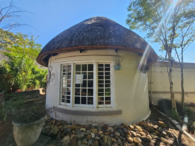 A unique circular structure featuring a traditional thatch roof, white exterior walls, and a bay window with security bars, set within a landscaped garden.