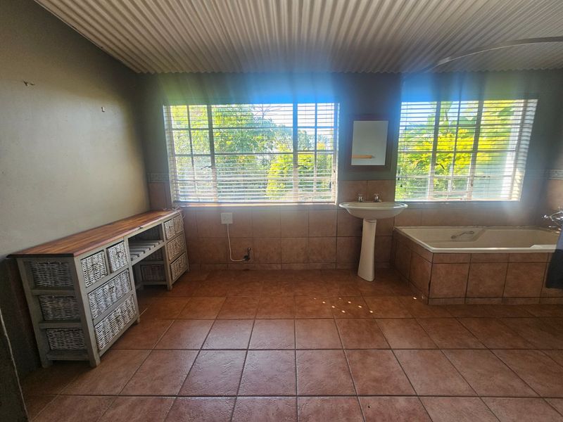 A spacious bathroom featuring a built-in bathtub, pedestal basin, tiled flooring, and large windows with venetian blinds, offering a functional layout with potential for modernization.
