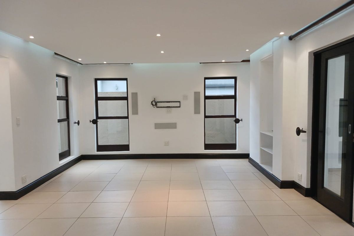A bright, empty living room featuring neutral tiled flooring, recessed downlights, built-in shelving, and modern black-framed windows and doors.