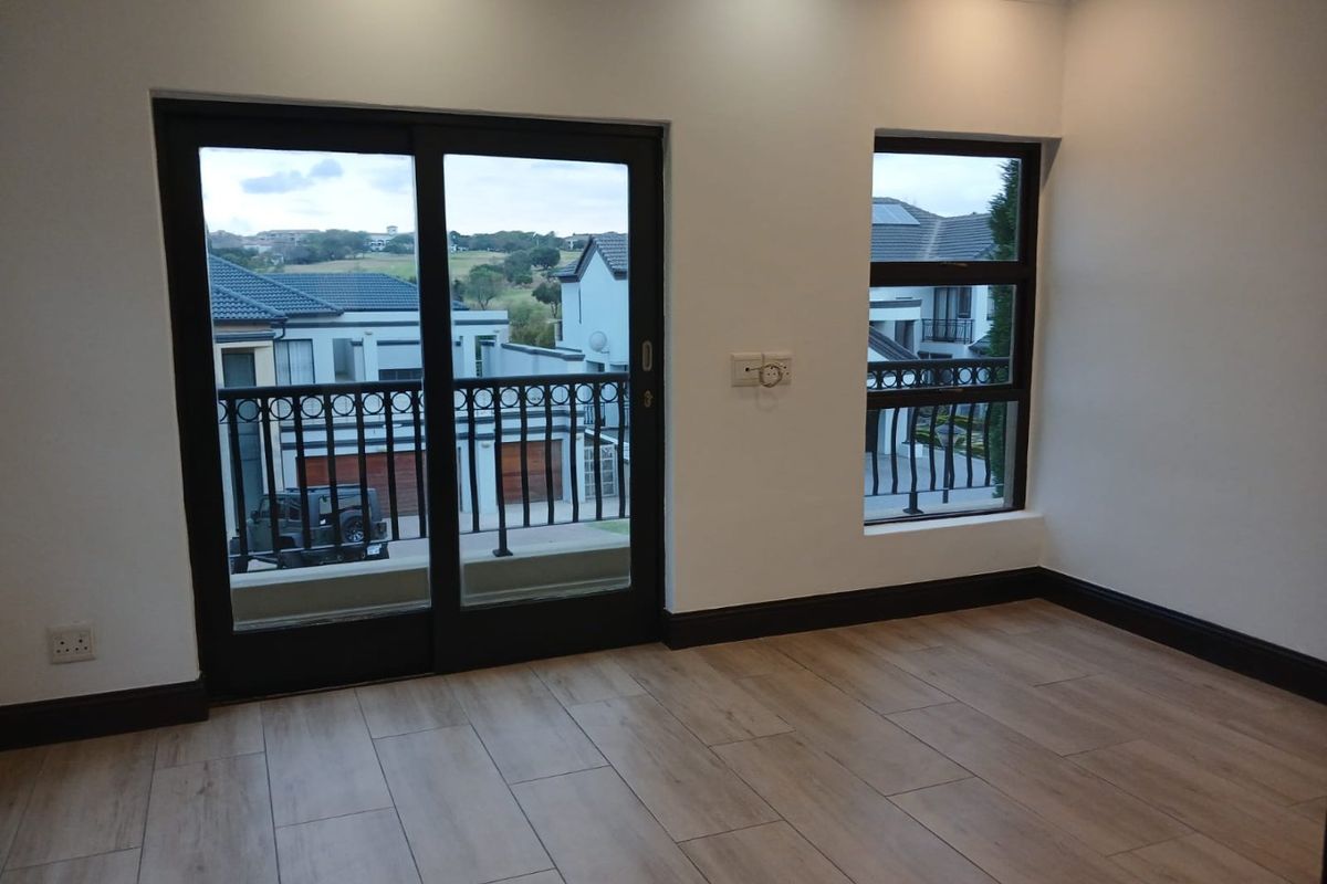 A bright, empty bedroom featuring modern wood-look tile flooring, a sliding glass door leading to a balcony, and large windows with neighbourhood views.