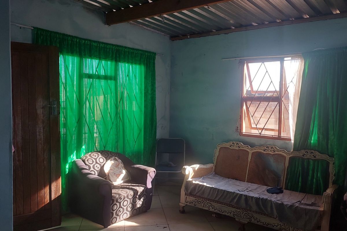 A living room featuring tiled flooring, security burglar bars on windows, and an exposed corrugated metal ceiling, requiring cosmetic updates to the wall finishes.