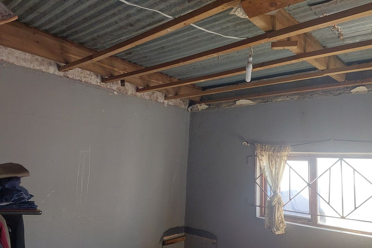 A bedroom space currently undergoing renovation, featuring exposed timber ceiling joists, corrugated metal roofing, and a window with security burglar bars.