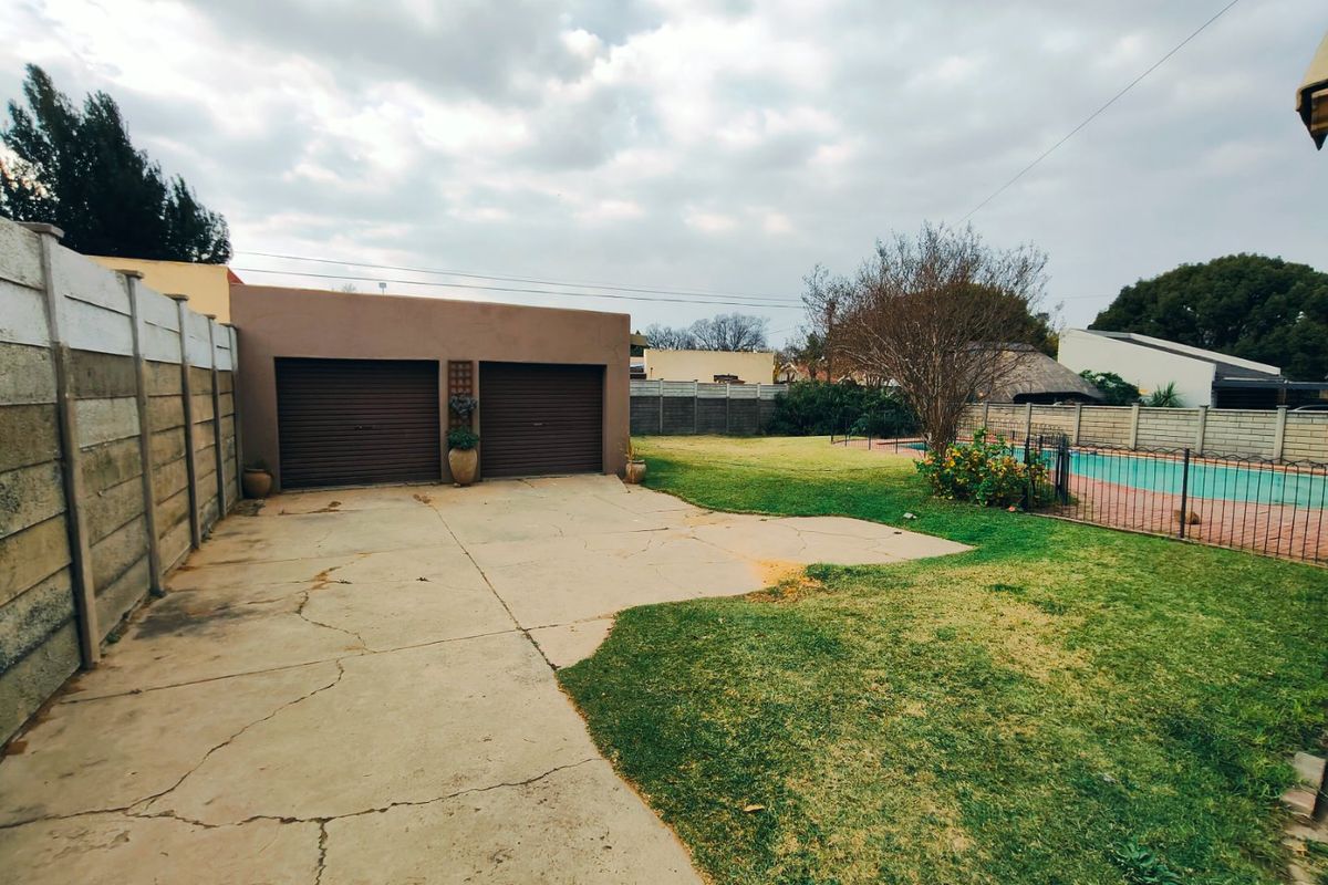 Rear exterior view featuring a double garage, concrete driveway with visible cracking, a lawn area, and a fenced swimming pool.