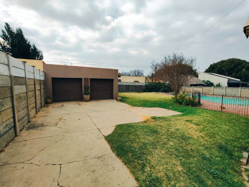 Rear exterior view featuring a double garage, concrete driveway with visible cracking, a lawn area, and a fenced swimming pool.