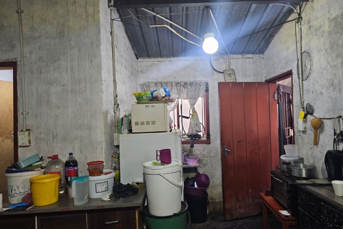 A kitchen area featuring exposed ceiling beams, visible electrical conduit, and a window with security bars, requiring significant renovation and wall surface repairs.