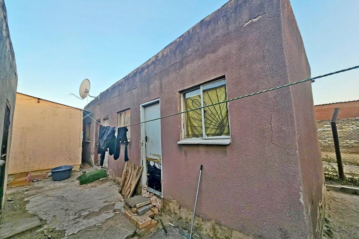 Rear exterior view of the property showing weathered plaster walls, sliding windows with security bars, and concrete paving.