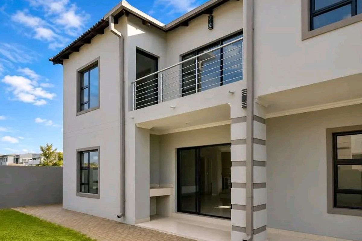 Rear view of a modern double-story home featuring a paved patio with a built-in braai, sliding glass doors, and an upper-level balcony with stainless steel railings.