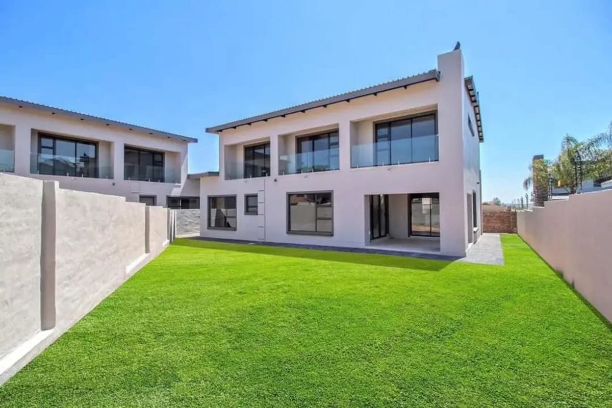 Rear view of a modern two-story house featuring a paved patio, glass balcony railings, large sliding doors, and a well-maintained lawn.