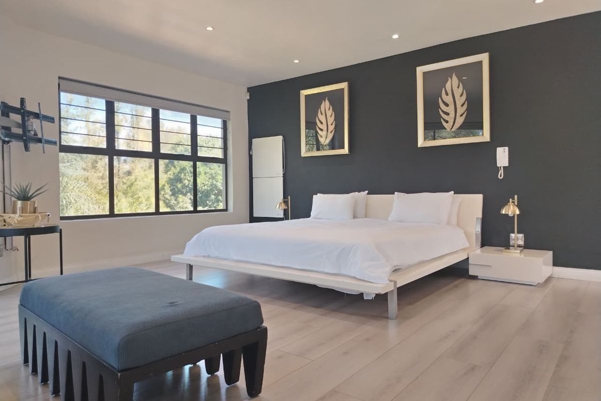 Spacious, modern bedroom featuring light laminate flooring, a large window providing natural light, and a dark feature wall with recessed downlighting.