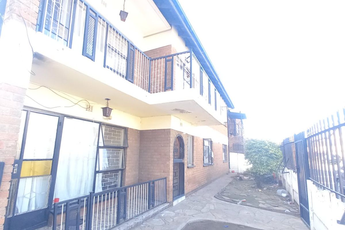 Rear exterior view of a two-story brick house featuring a balcony, security gates, and a paved walkway leading to a small garden area.
