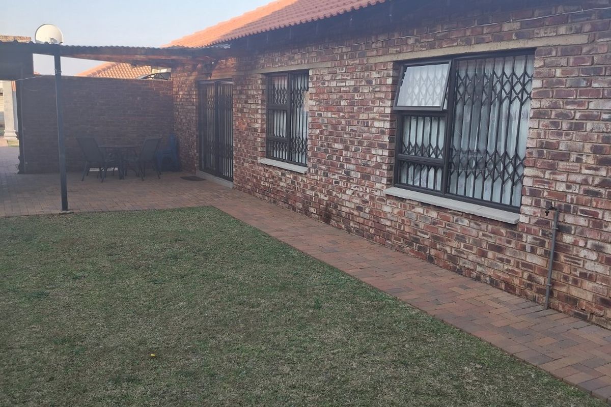 Rear view of a face brick townhouse featuring a tiled roof, security gates on windows, a paved walkway, and a small covered patio area.
