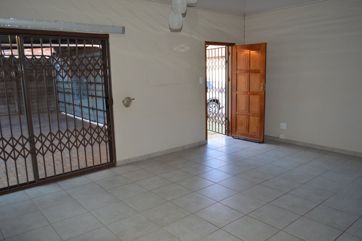 An empty living area featuring neutral tiled flooring, a timber entrance door, and sliding security gates leading to the exterior.