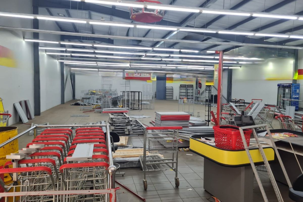 Large open-plan commercial retail space featuring exposed steel roof trusses, tiled flooring, and integrated checkout counters, currently in a cluttered state.