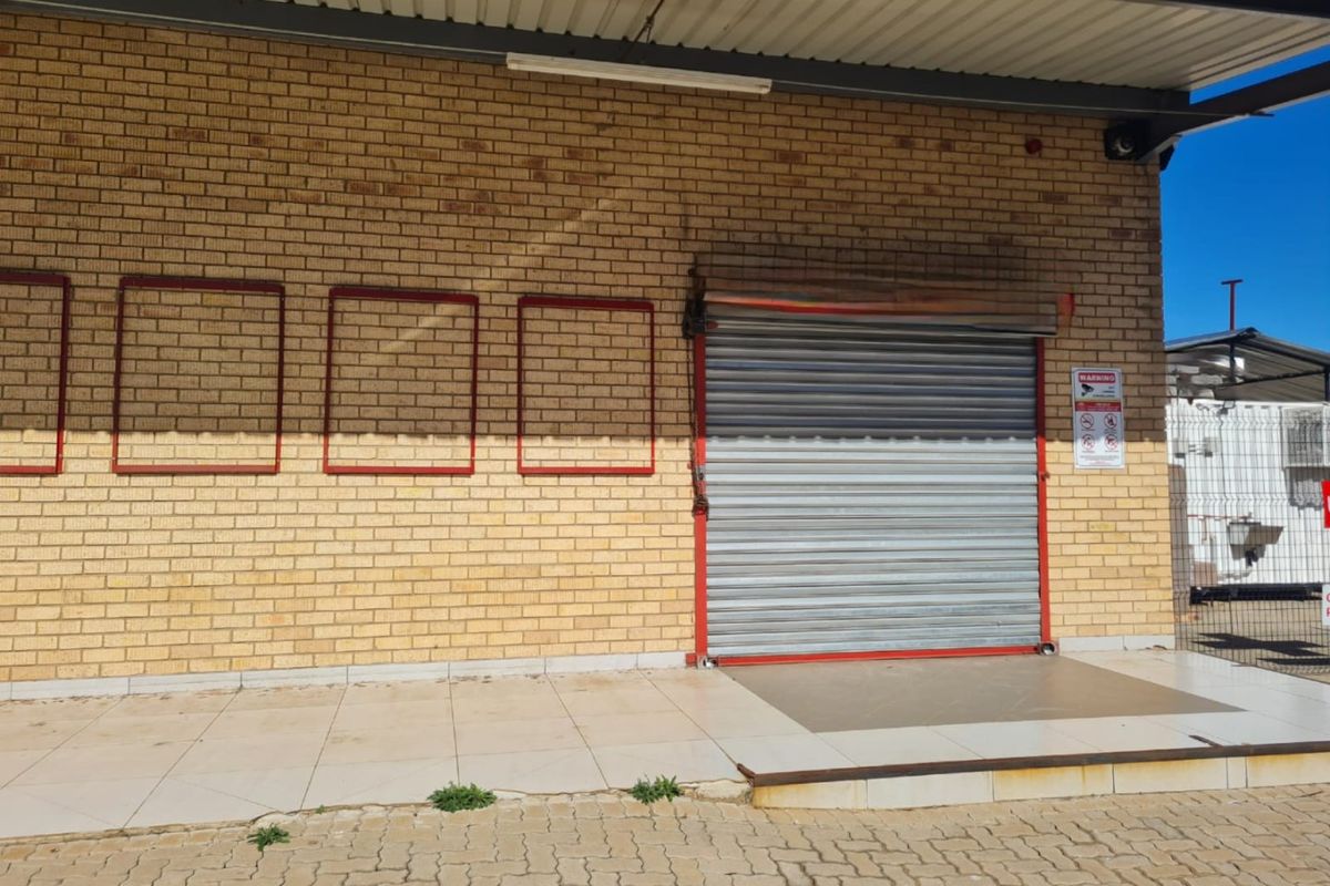 Commercial building exterior featuring a brick facade, a secure roller shutter door, and a tiled entrance area with paved driveway access.