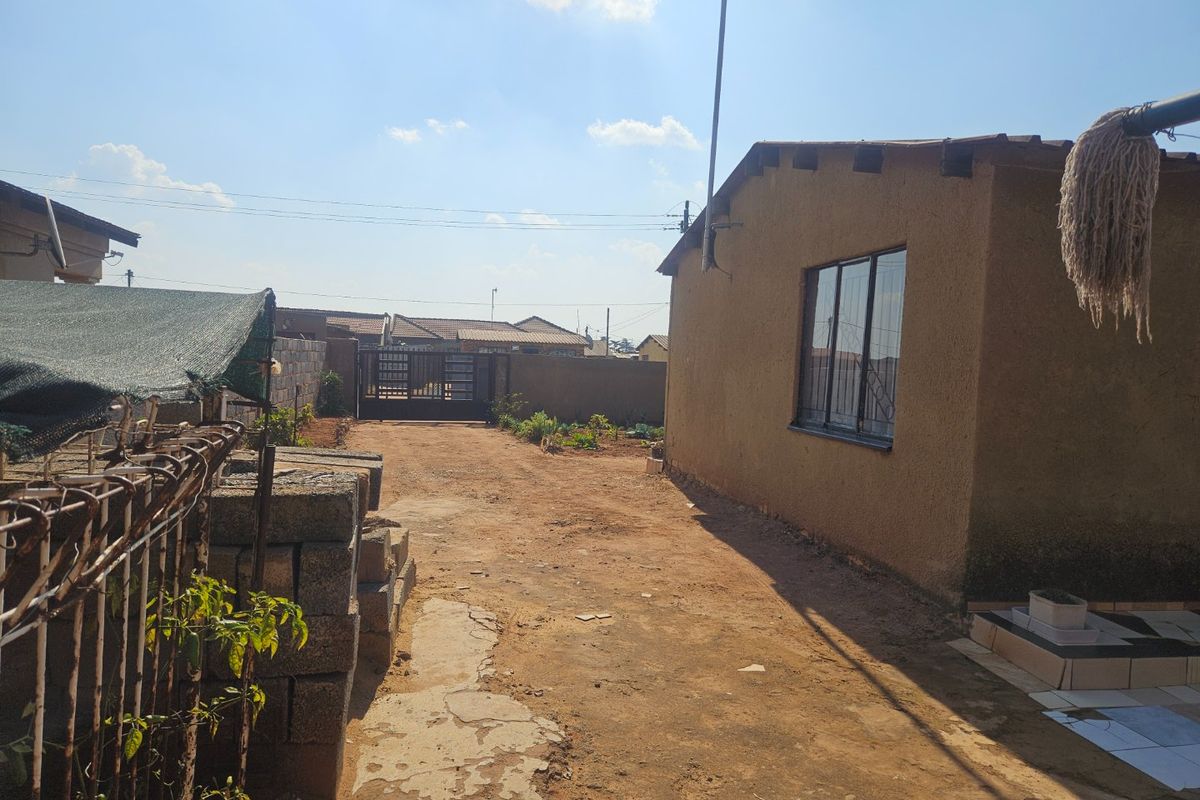 Rear exterior view showing a side wall, unpaved driveway area, boundary wall with metal gate, and a small tiled patio section.